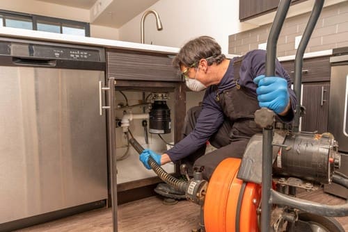 Plumber with motorized drain snake cleaning out a drain under a sink